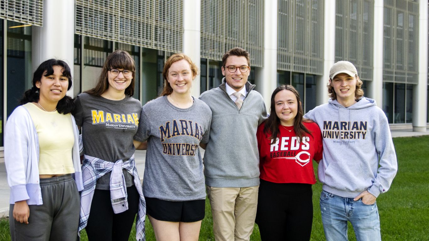 A group of six students poses outside a building, showcasing their Marian University apparel.