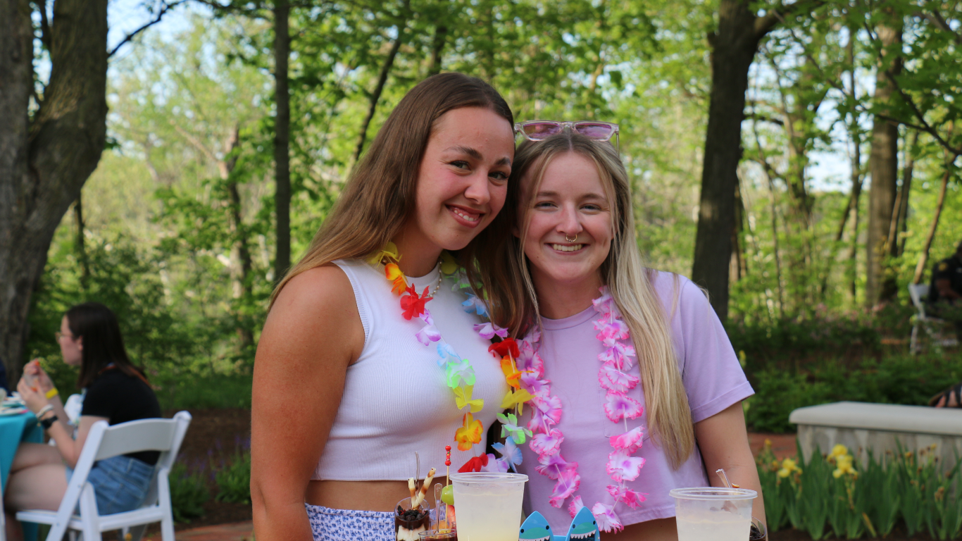 Two young women smiling and wearing colorful leis at an outdoor gathering, with drinks in front of them.