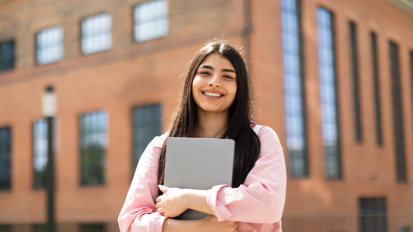 A smiling young woman holding a laptop in front of a modern urban building.