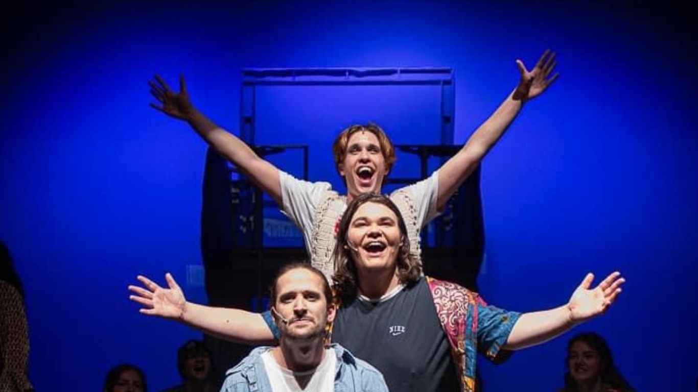 Three performers on stage striking a joyful pose against a blue backdrop, showcasing an expressive and vibrant moment from a theatrical performance.