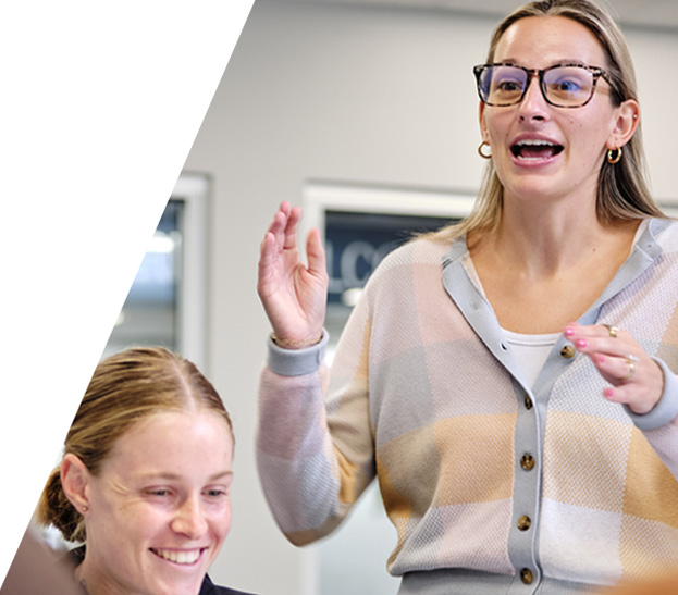 Woman wearing glasses speaks and gestures while standing in an office meeting, with a smiling colleague seated nearby.