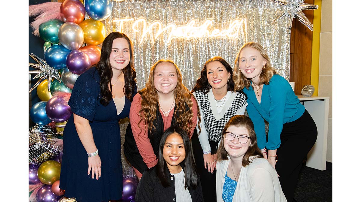 A group of six women posing together in front of a sparkling backdrop with colorful balloons and a illuminated sign.