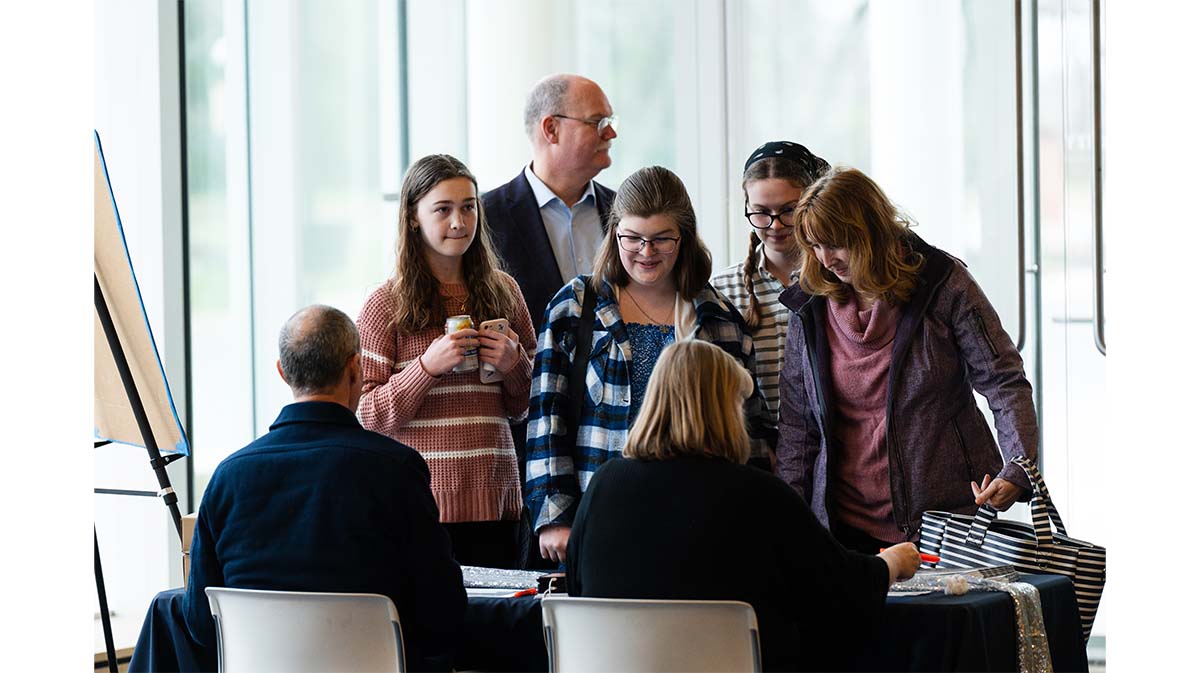 A group of people engaging with a table, likely at an event, with one person seated and interacting with a few attendees.