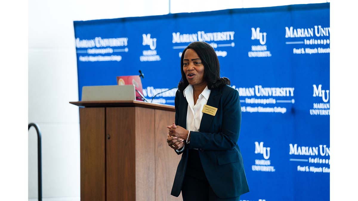 A speaker presenting at a podium in front of a Marian University backdrop during a university event.