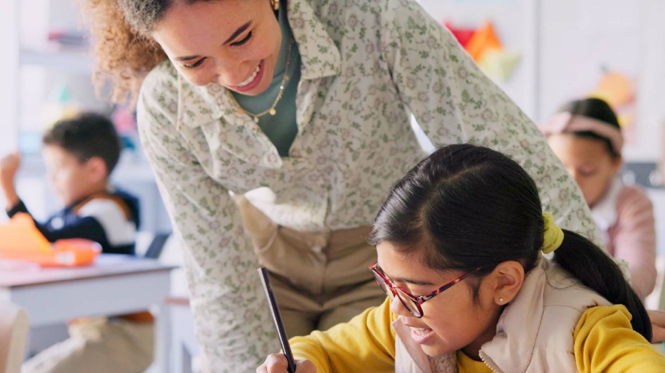 A teacher assists a young student with her writing in a classroom setting. The scene captures a supportive learning environment with other students engaged in the background.