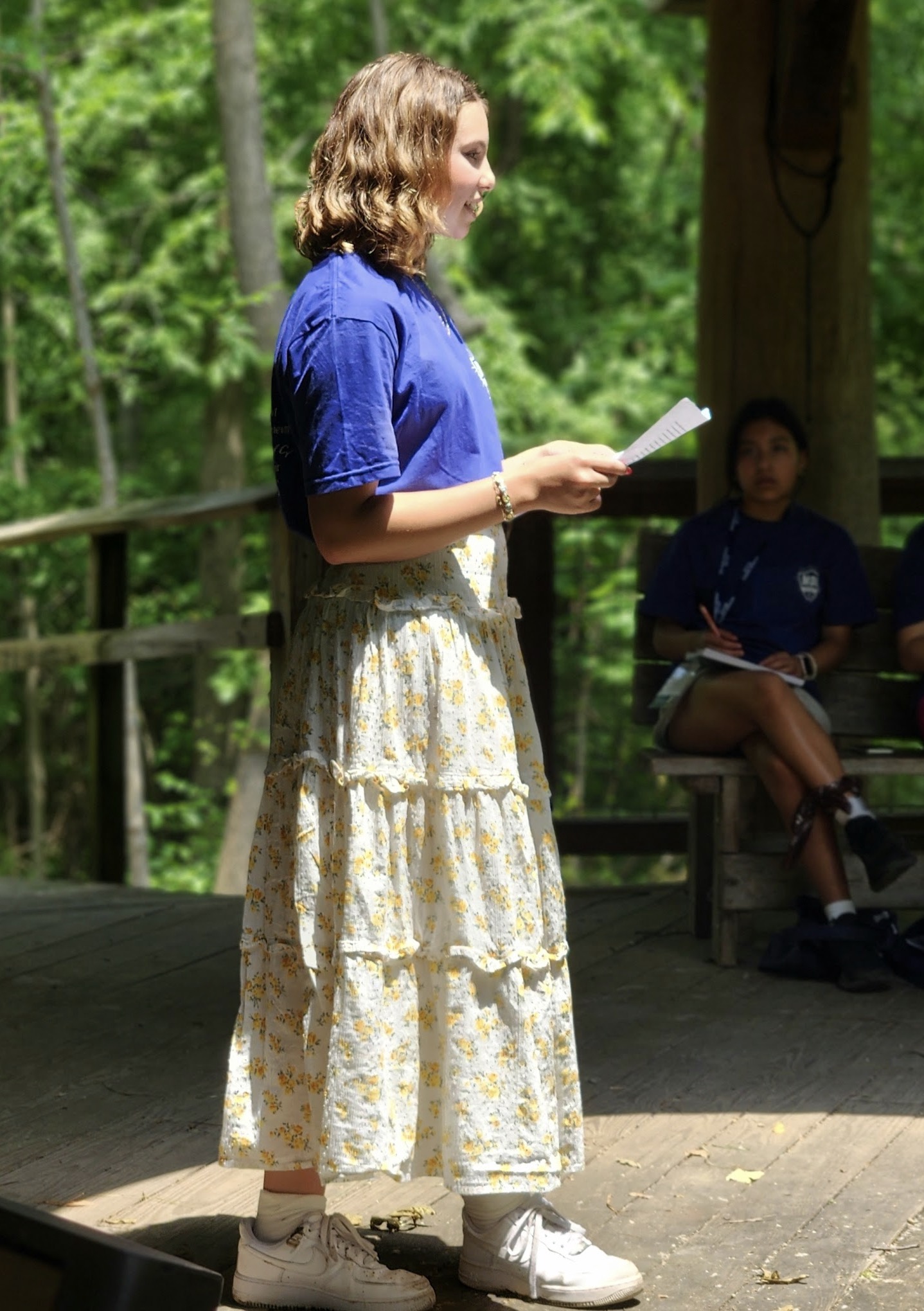 A young girl in a blue shirt and floral skirt stands on a stage, reading from a paper, while others sit in the background.