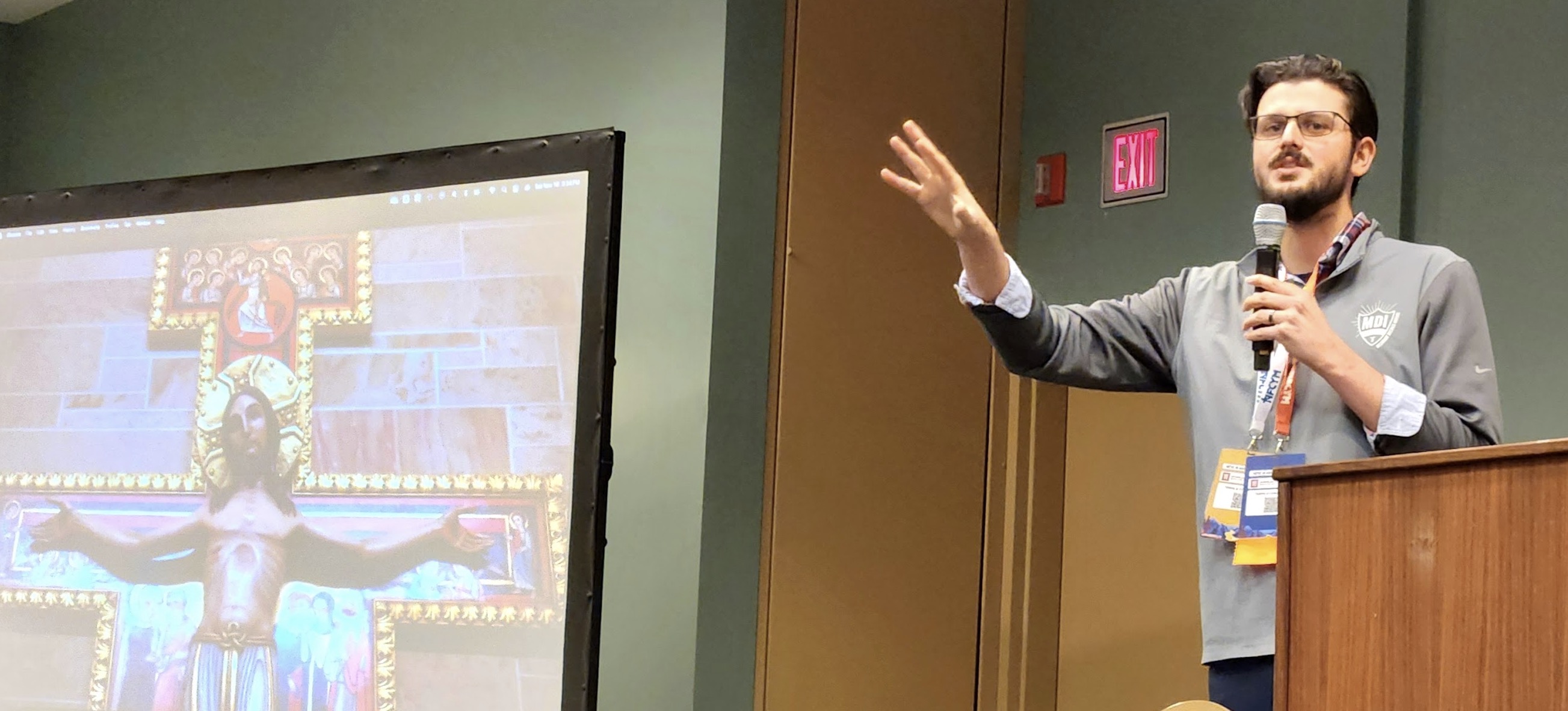 A speaker presenting at a conference with a large image of a crucifixion artwork displayed behind him.