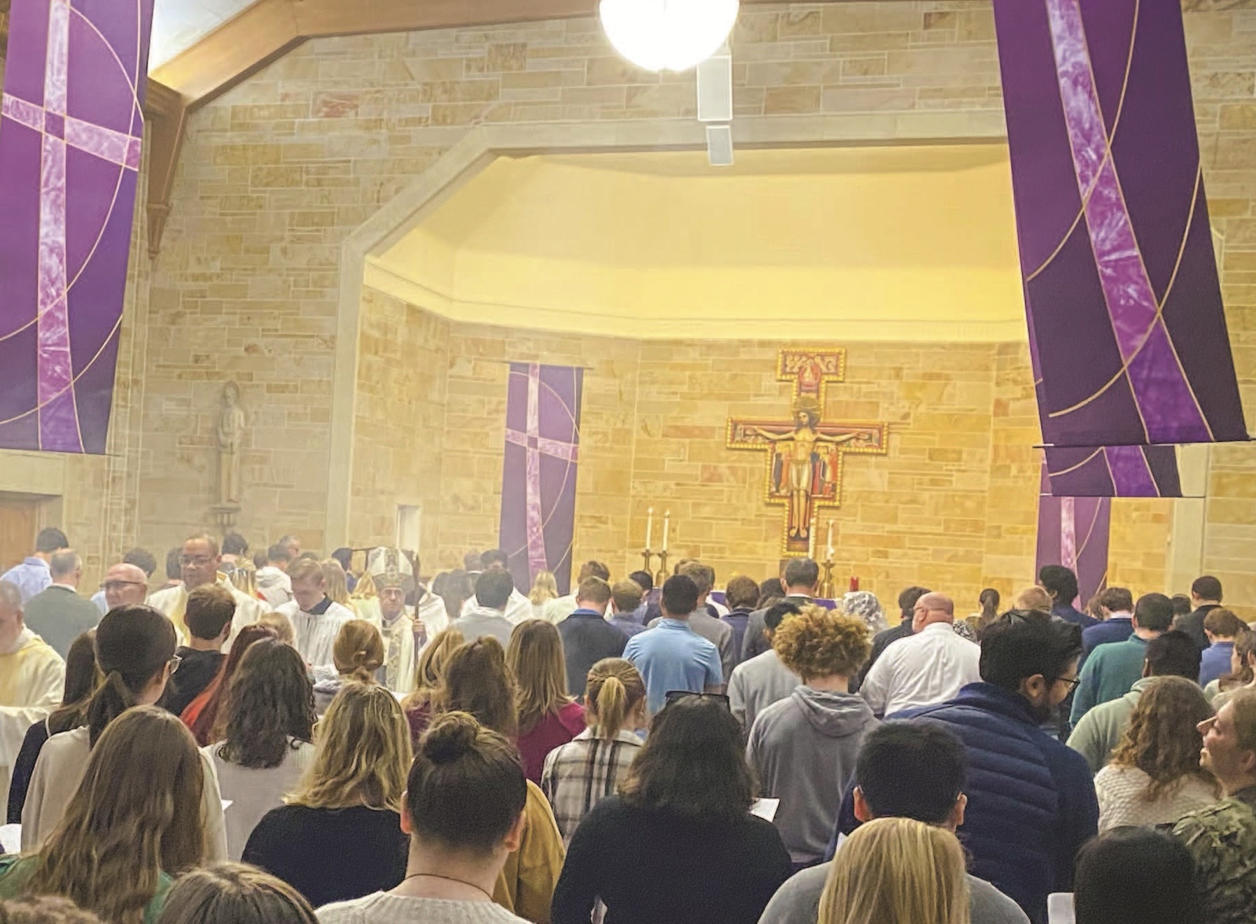 Congregation gathered for a church service, facing an altar with a crucifix and purple banners.