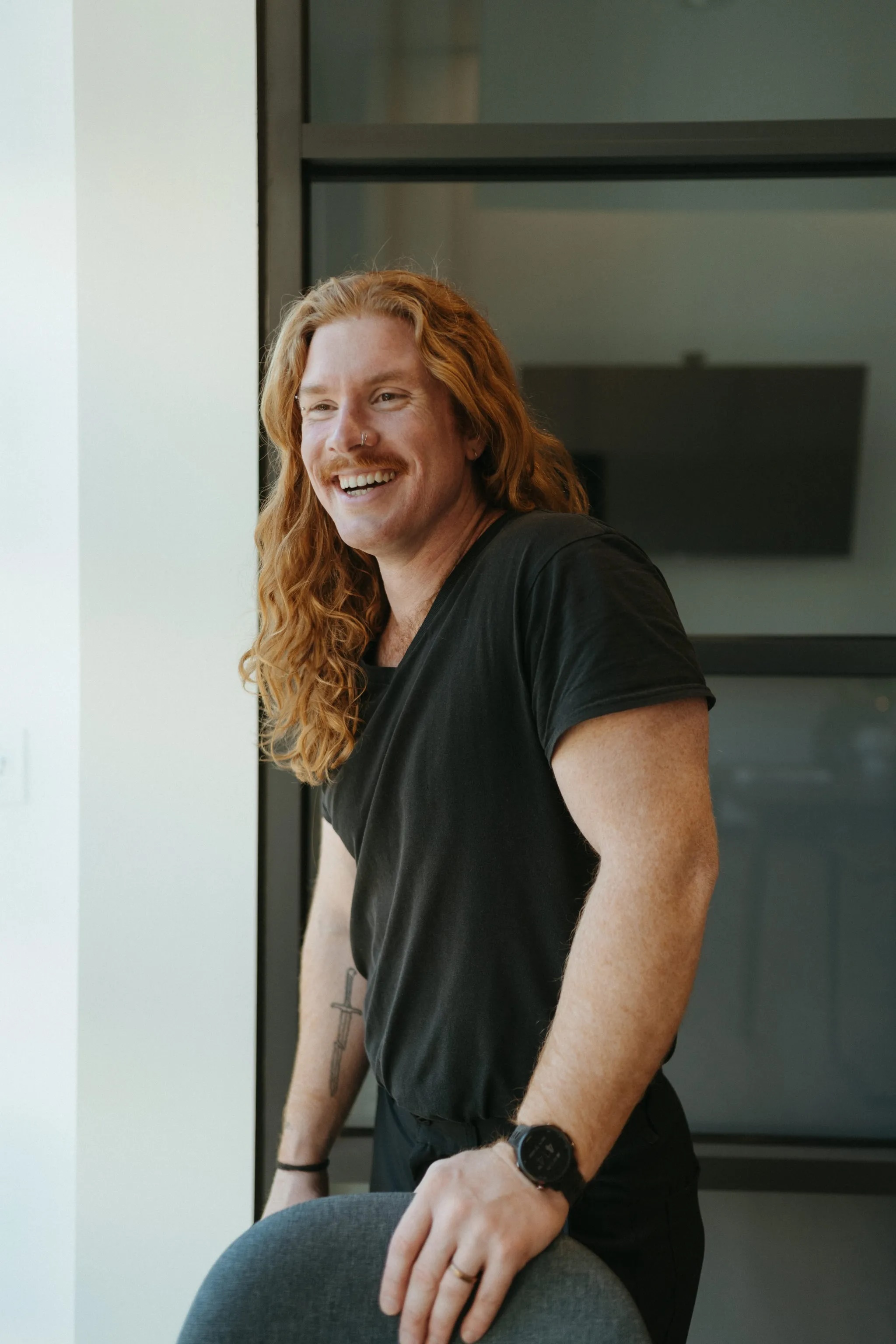 A smiling man with long, wavy red hair and a mustache poses in a relaxed manner, wearing a black shirt and standing next to a chair in a modern office setting.