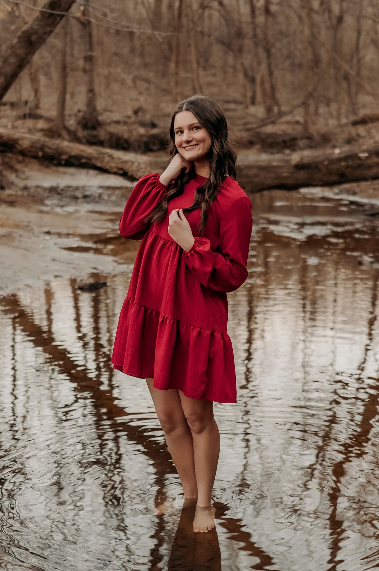 A young woman stands in a shallow stream wearing a red dress, surrounded by trees in a natural setting.