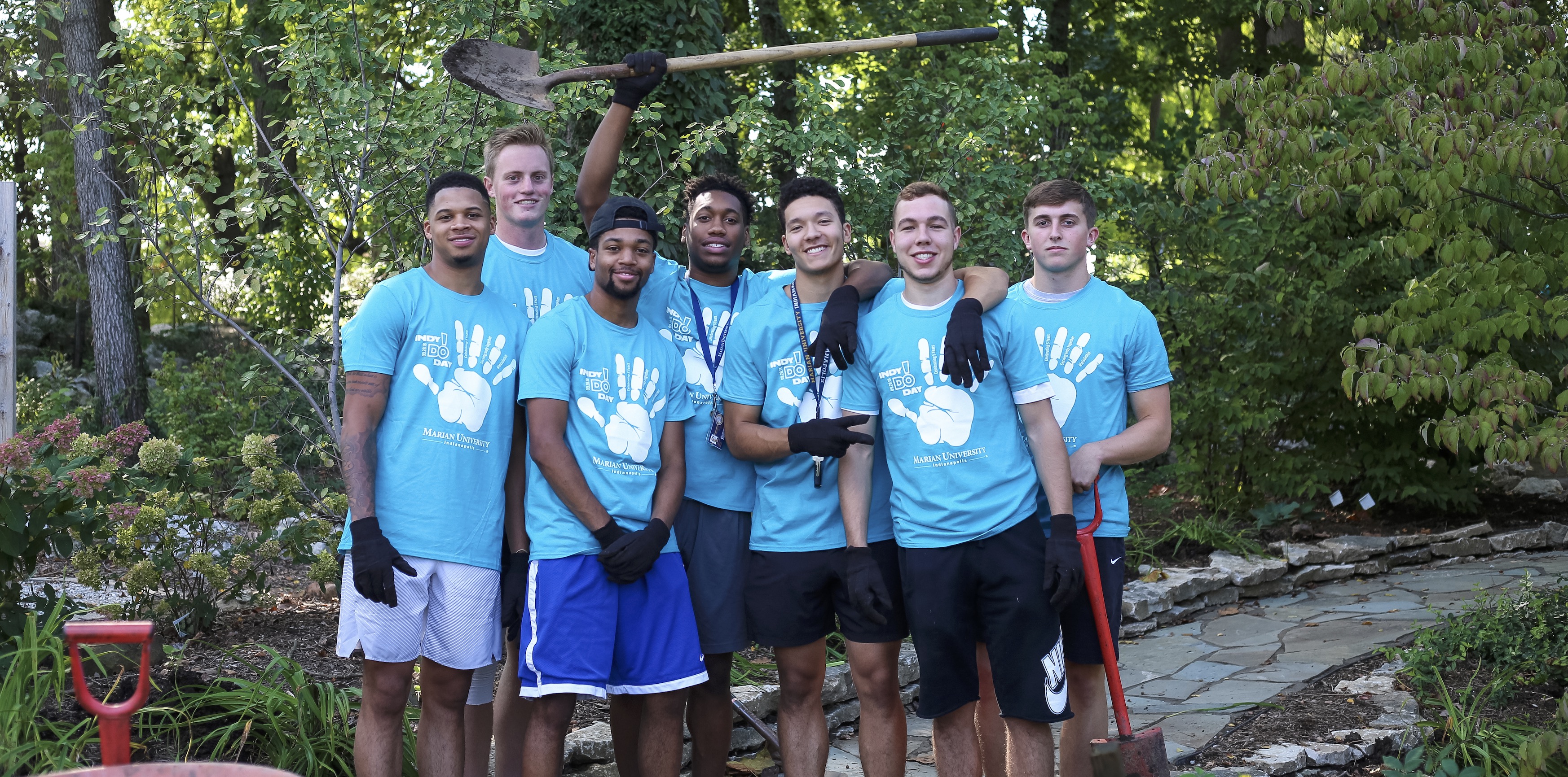 Group of student volunteers in matching blue shirts and work gloves posing in a garden, holding shovels.