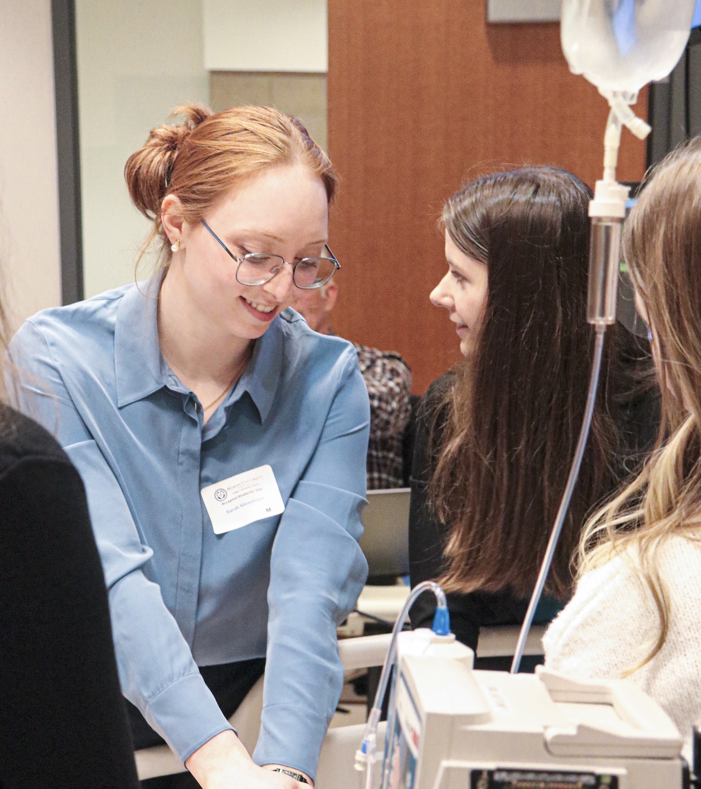 Nursing student demonstrating IV equipment while talking with classmates in a clinical skills lab.