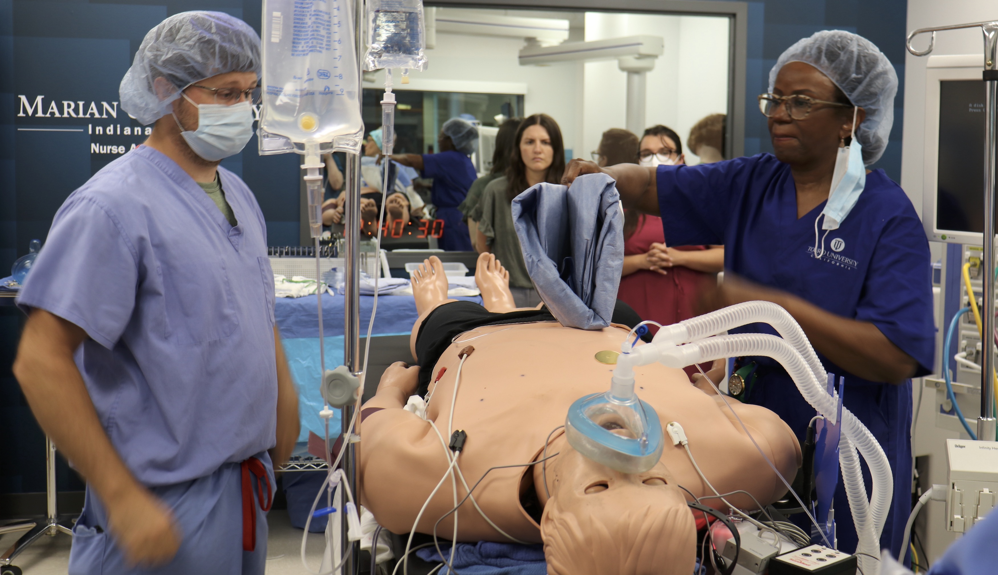Nursing students practice a medical procedure on a simulation mannequin in a lab, with IV equipment and monitoring devices attached.