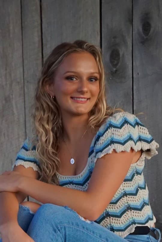A young woman with long, wavy hair poses in front of a wooden backdrop, wearing a stylish crocheted top and jeans.