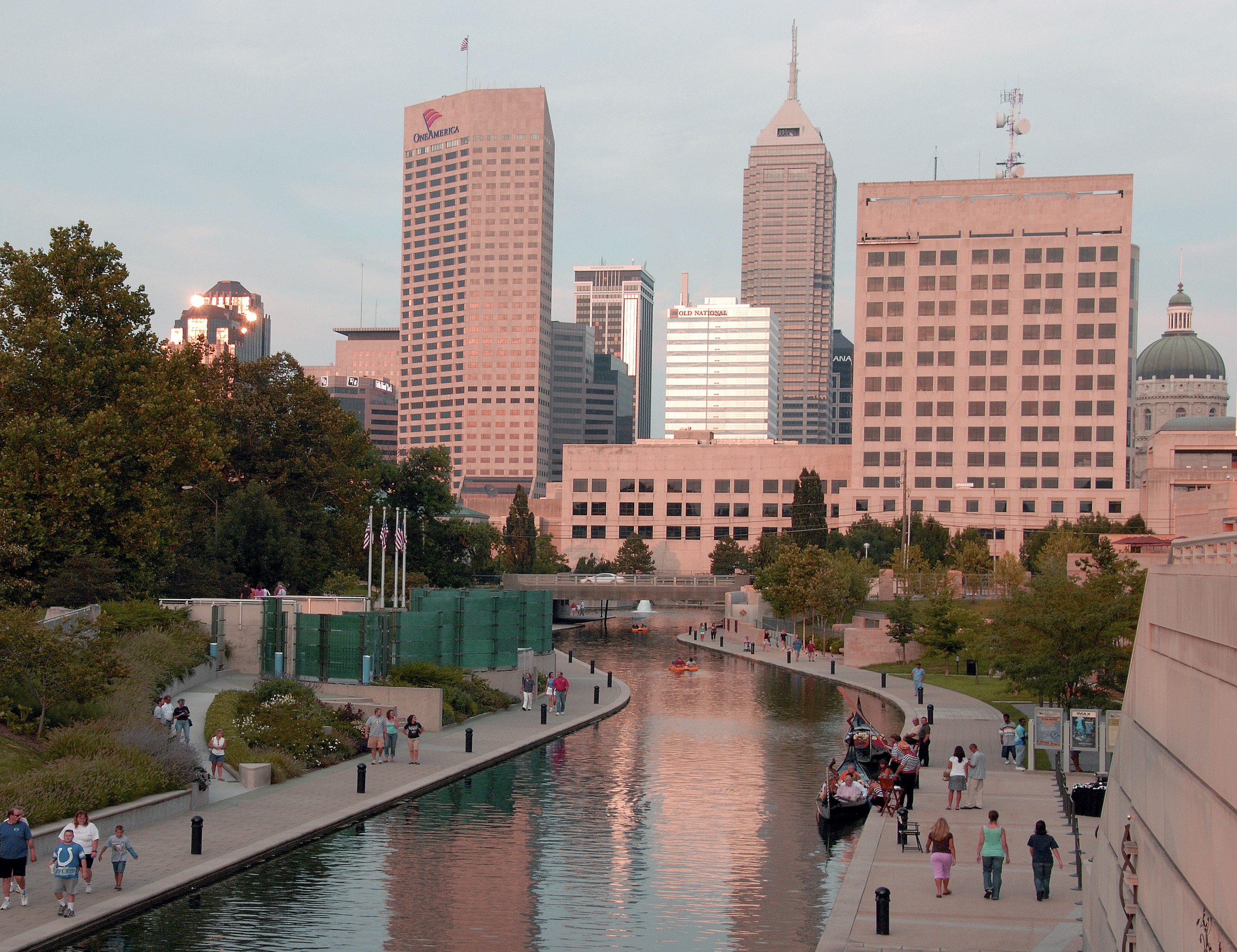 Downtown skyline at sunset behind a canal walkway with people strolling and boats on the water.