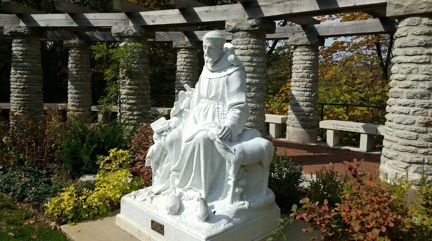 White stone statue of a robed monk seated under a pergola in a garden, with stone columns and memorial benches in the background.