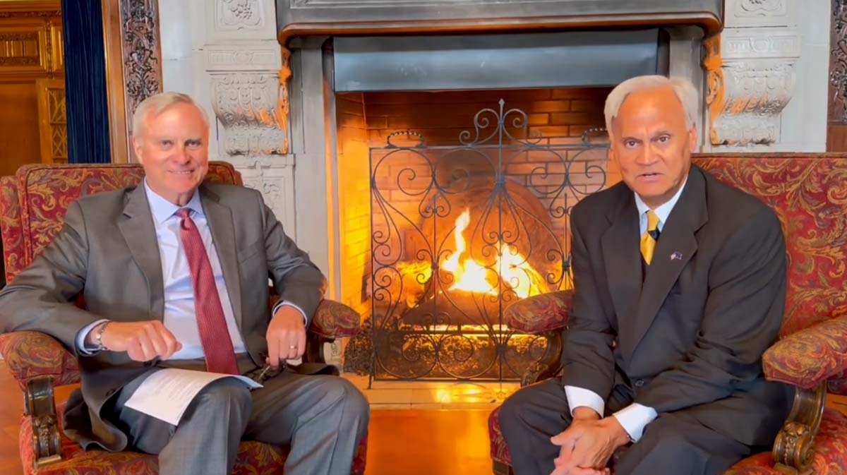Two men seated in ornate chairs in front of a fireplace, engaged in conversation.