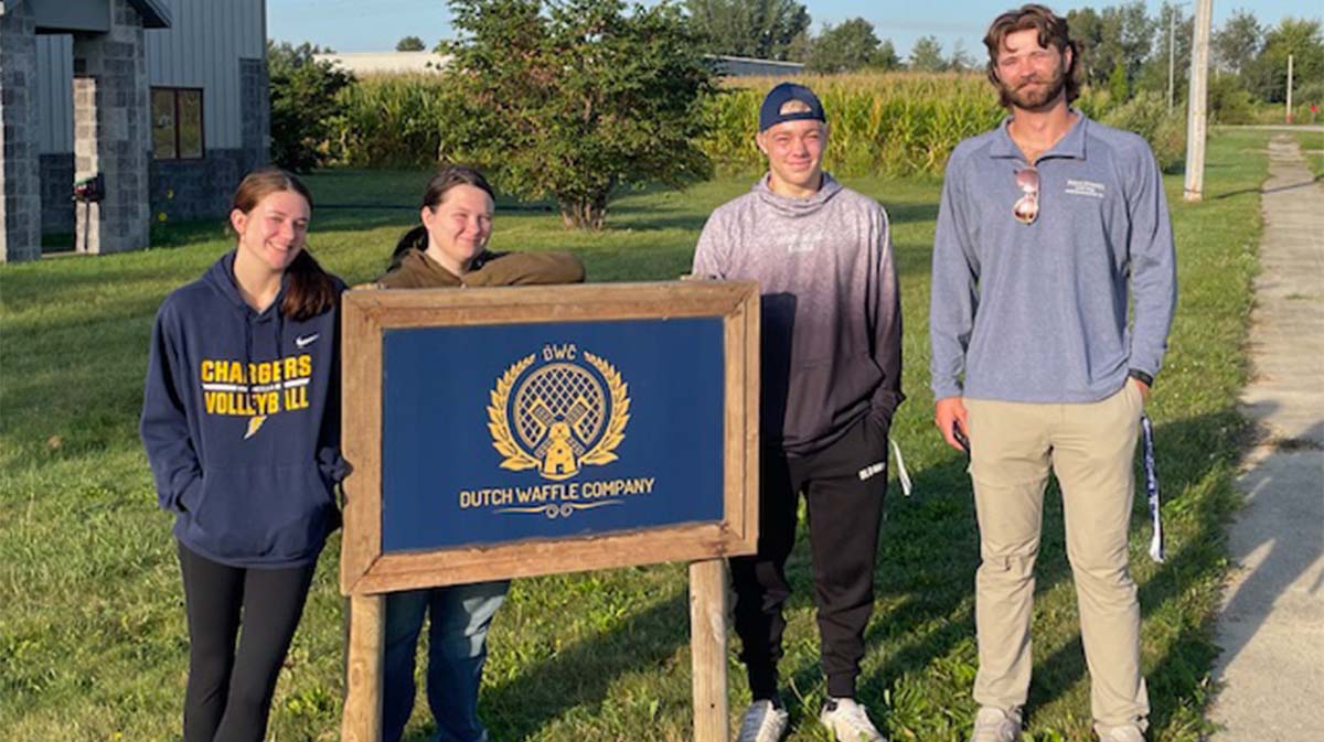 Group of four people standing by the Dutch Waffle Company sign in a grassy area.