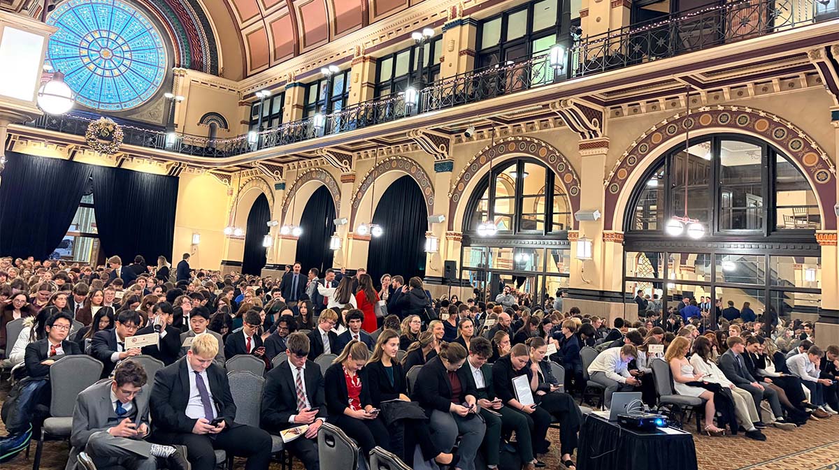 A packed auditorium filled with attendees in formal attire, engaged in conversation or using their phones, with ornate architecture and a large stained glass window in the background.