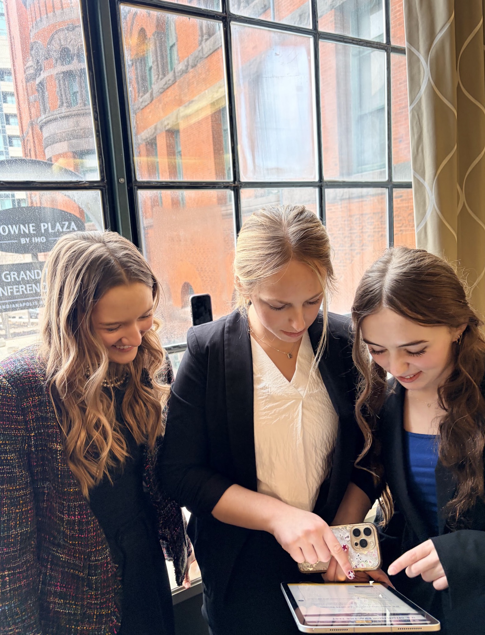 Three young women gathered by a window, collaboratively looking at a tablet.
