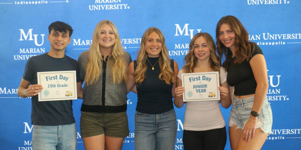 Five students pose in front of a Marian University Indianapolis step-and-repeat backdrop, with two holding “First Day” signs.