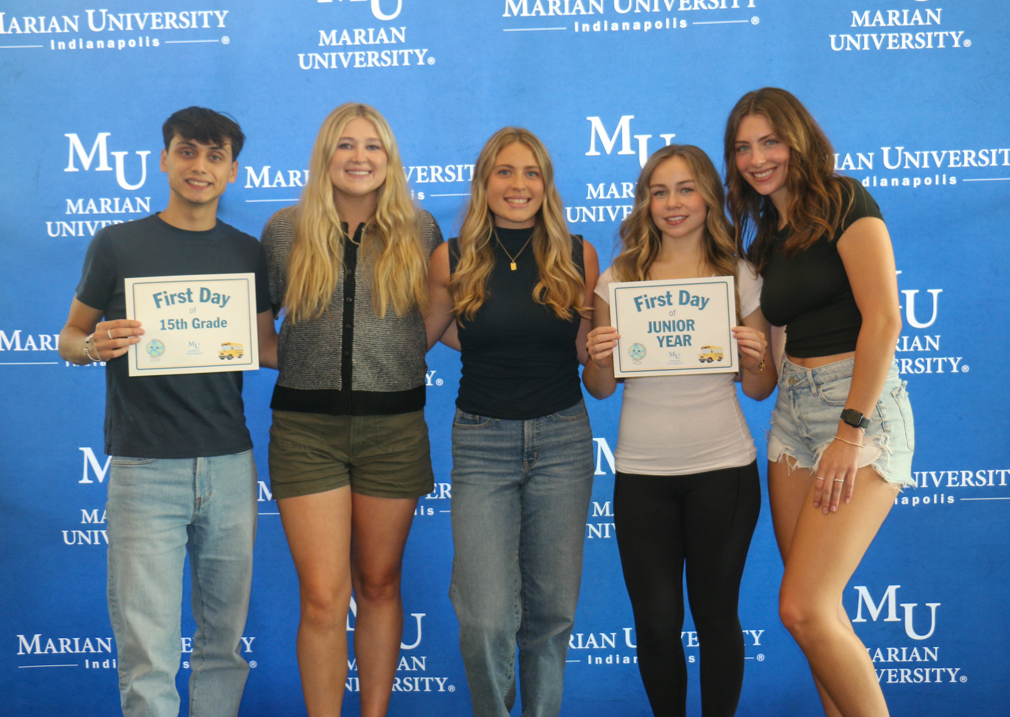 Five students pose in front of a Marian University Indianapolis step-and-repeat backdrop, with two holding “First Day” signs.