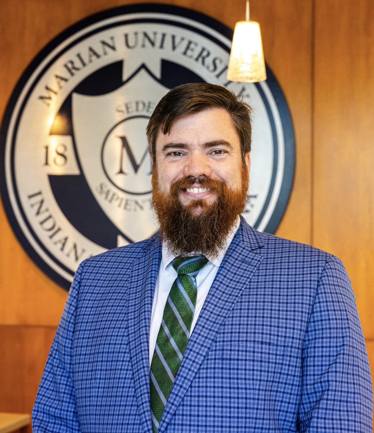 Smiling man in a blue checkered suit and green striped tie standing indoors in front of a Marian University seal.