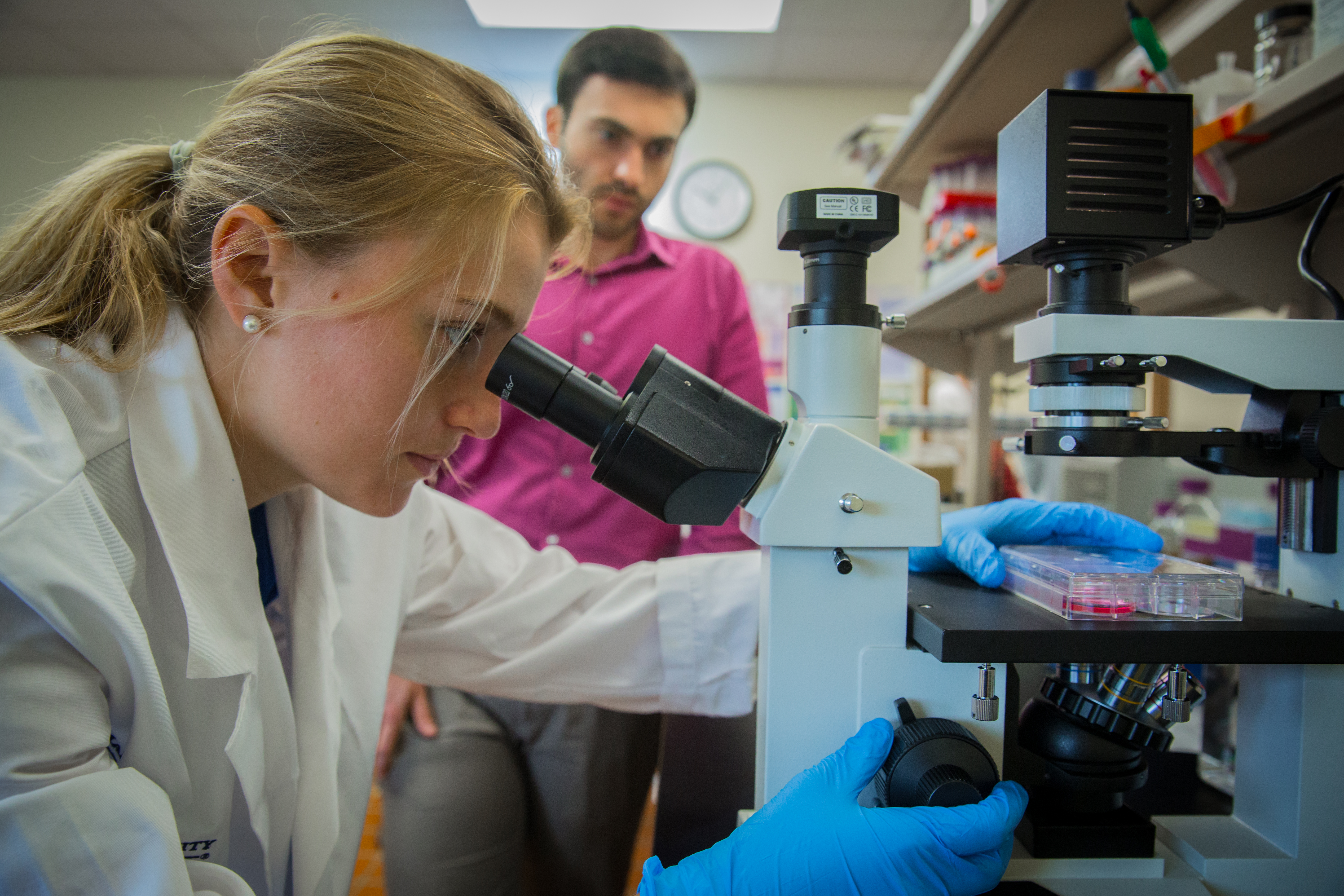 Researcher in a lab coat looks through a microscope while a colleague stands in the background near a lab bench.