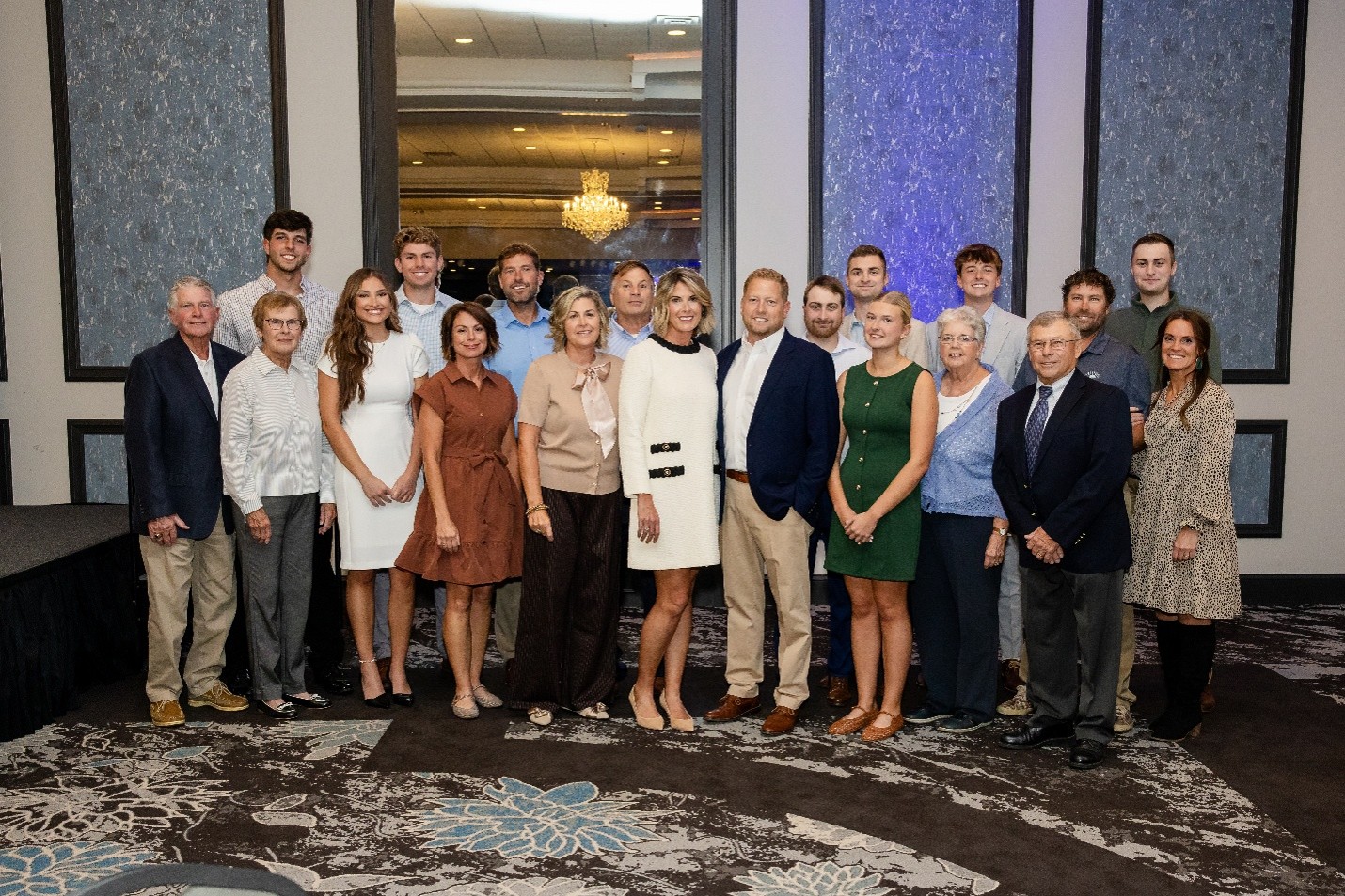 Large group of people posing for a formal photo in a hotel ballroom, with a chandelier and tall blue wall panels behind them.