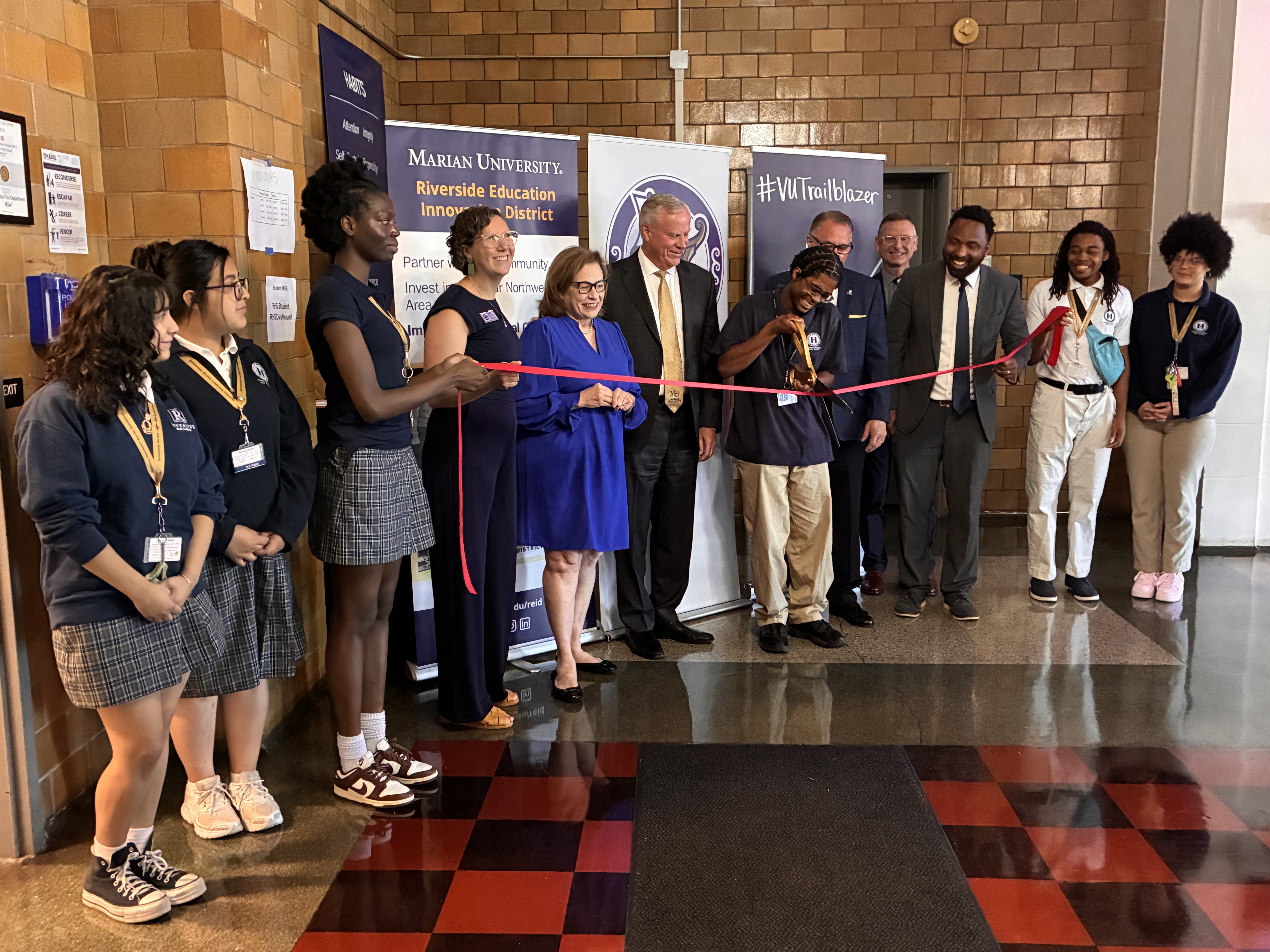 Group of students and staff at a ribbon-cutting ceremony, with a student cutting a red ribbon in front of banners for Marian University and #VUTrailblazer.