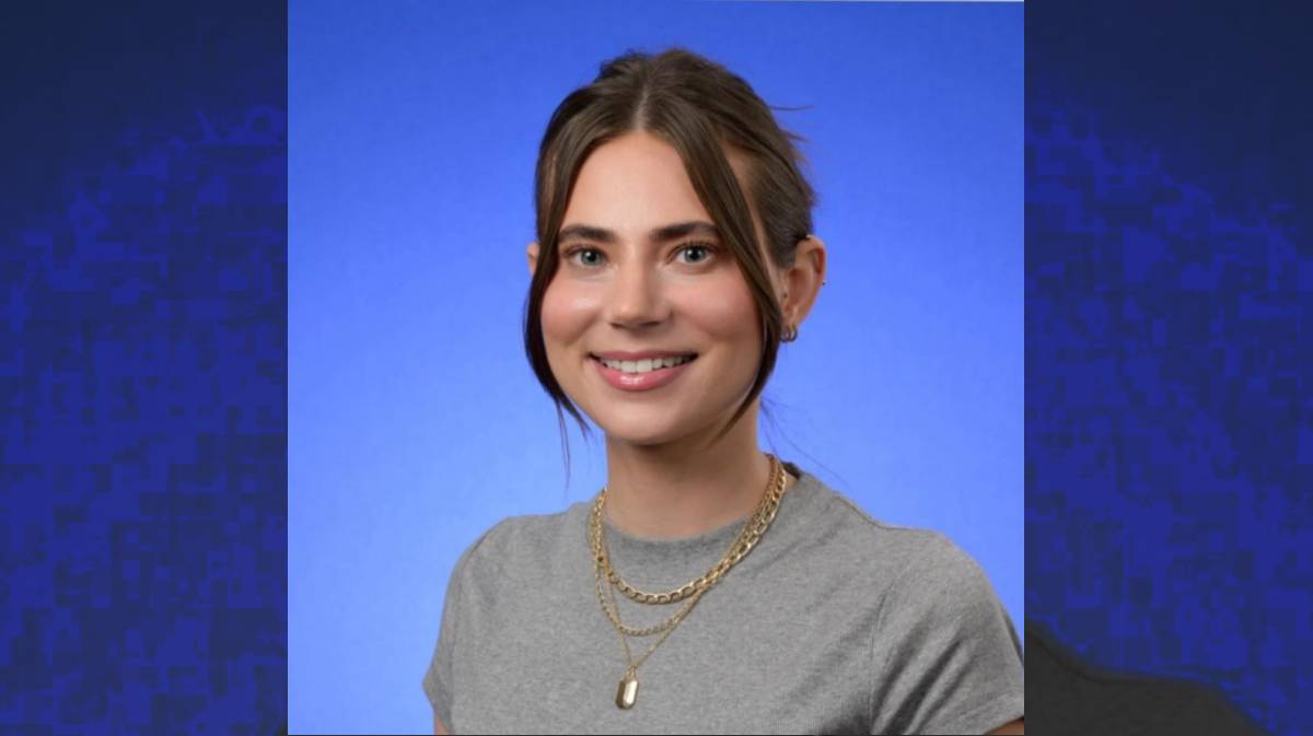 Smiling person in a gray T-shirt and layered gold necklaces against a blue background.