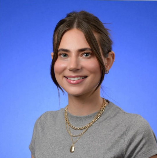 Smiling person in a gray T-shirt and layered gold necklaces against a blue background.