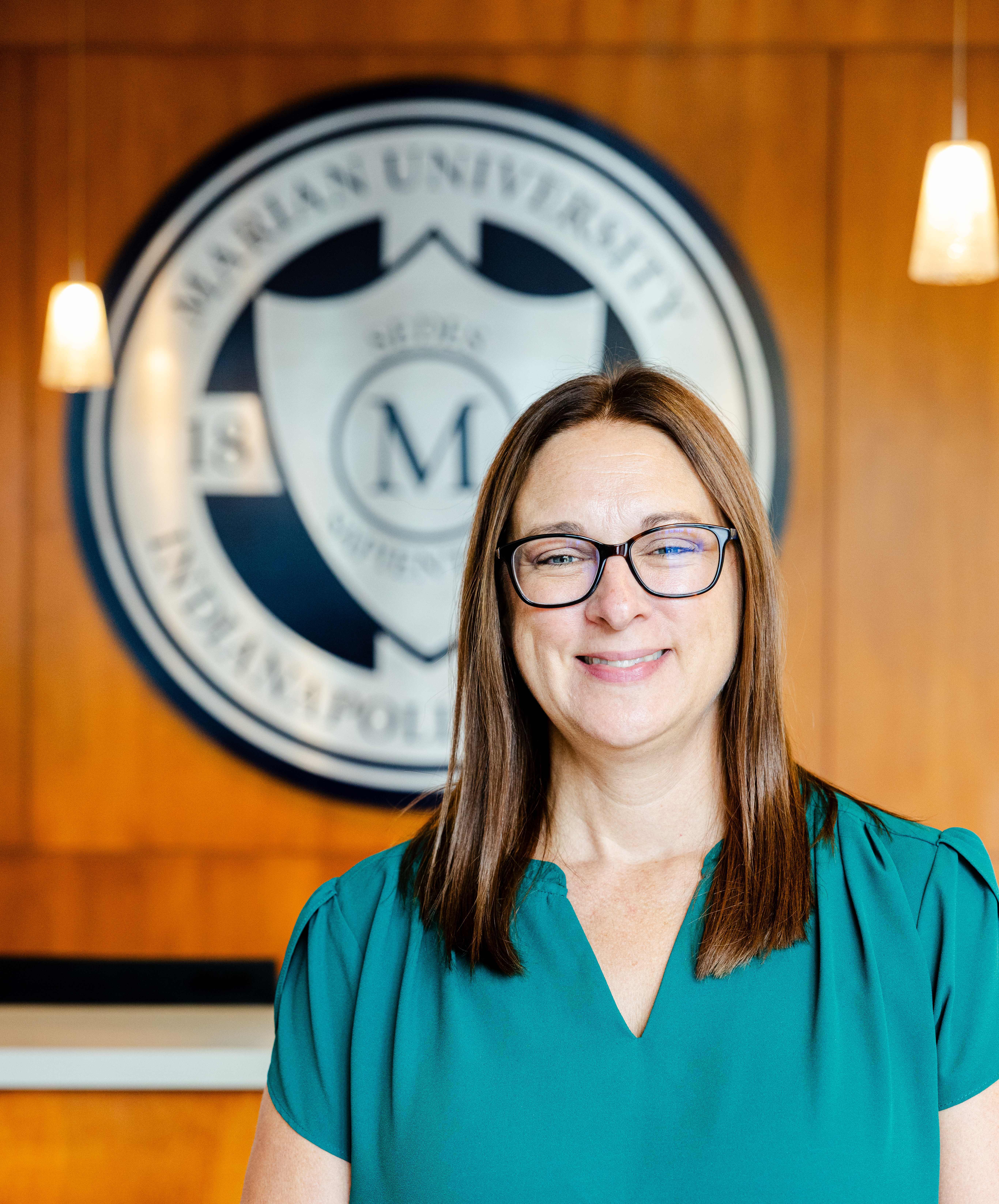 Smiling woman with glasses in a teal blouse standing indoors, with a blurred university seal in the background.