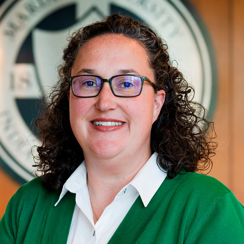 Head-and-shoulders portrait of a smiling woman with curly hair and glasses, wearing a green sweater over a white collared shirt, with a blurred circular emblem in the background.