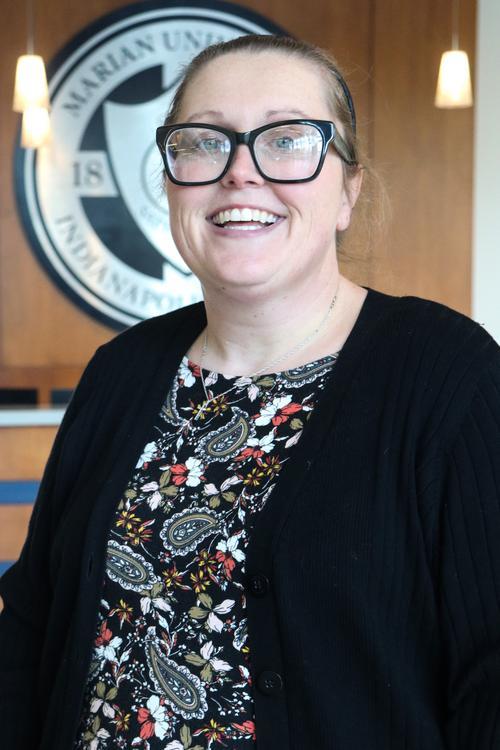 Smiling woman wearing large black glasses and a floral top, standing indoors with a blurred university seal in the background.