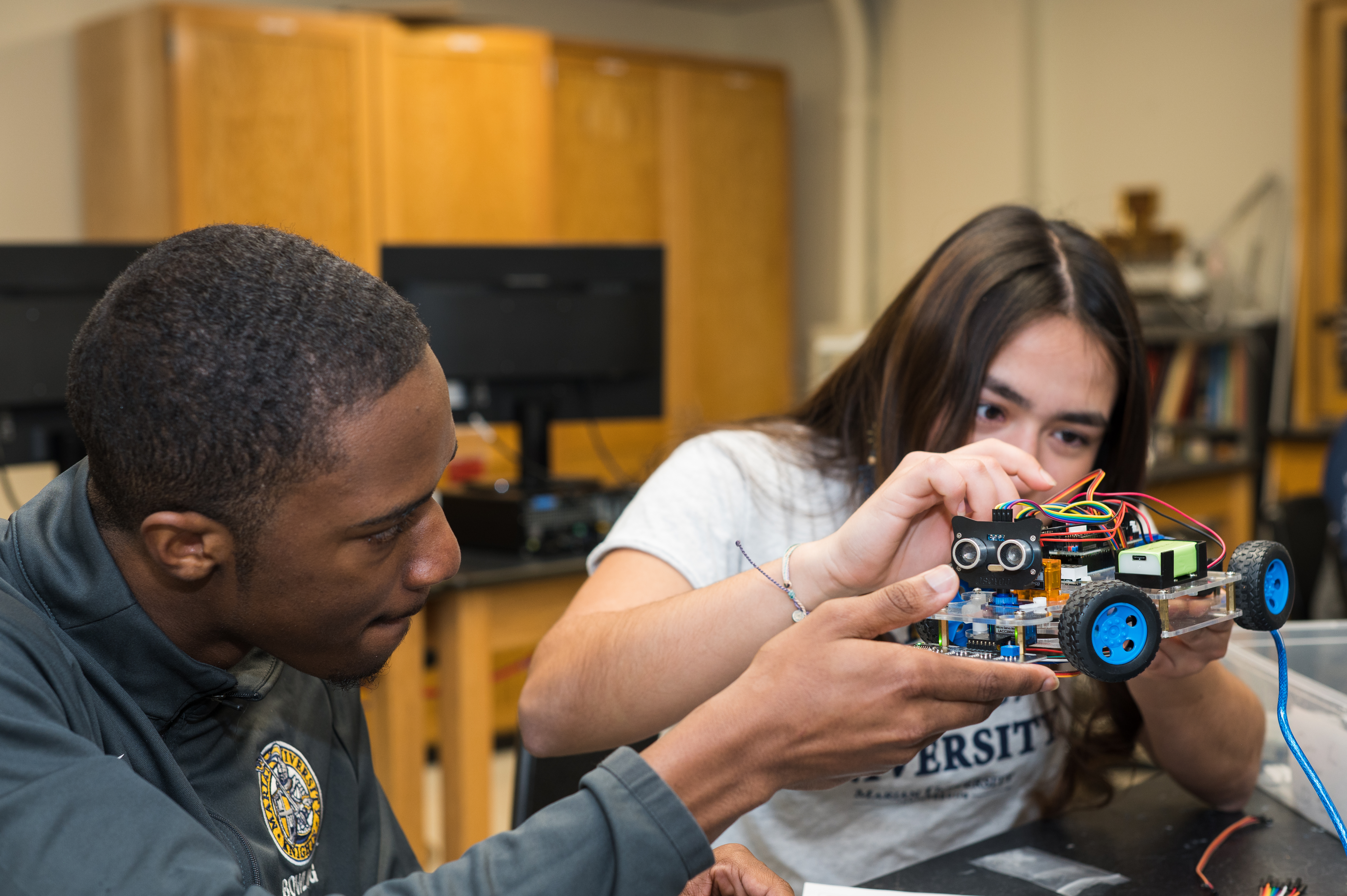 Two students work together on a small wheeled robot with exposed wiring and sensors in a classroom lab.