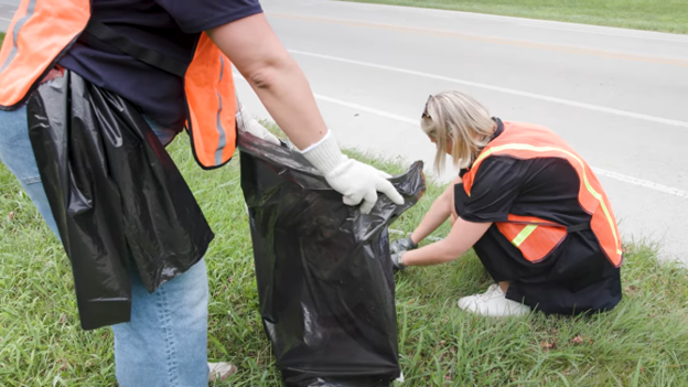 Two volunteers wearing orange safety vests collect litter into a large black trash bag along a roadside grassy area.