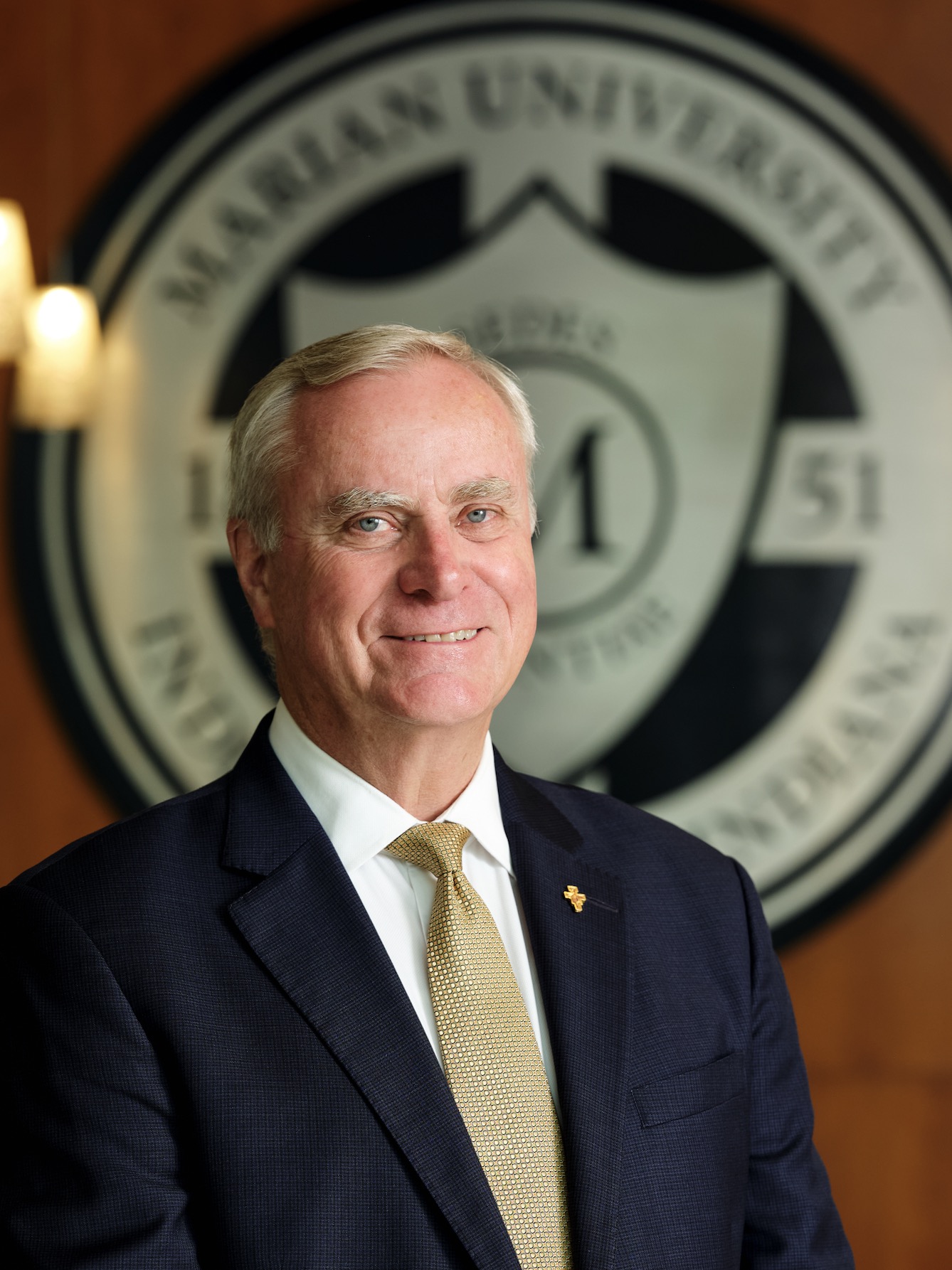 Portrait of an older man in a dark suit and gold tie, standing in front of a blurred circular emblem with candles to the side.