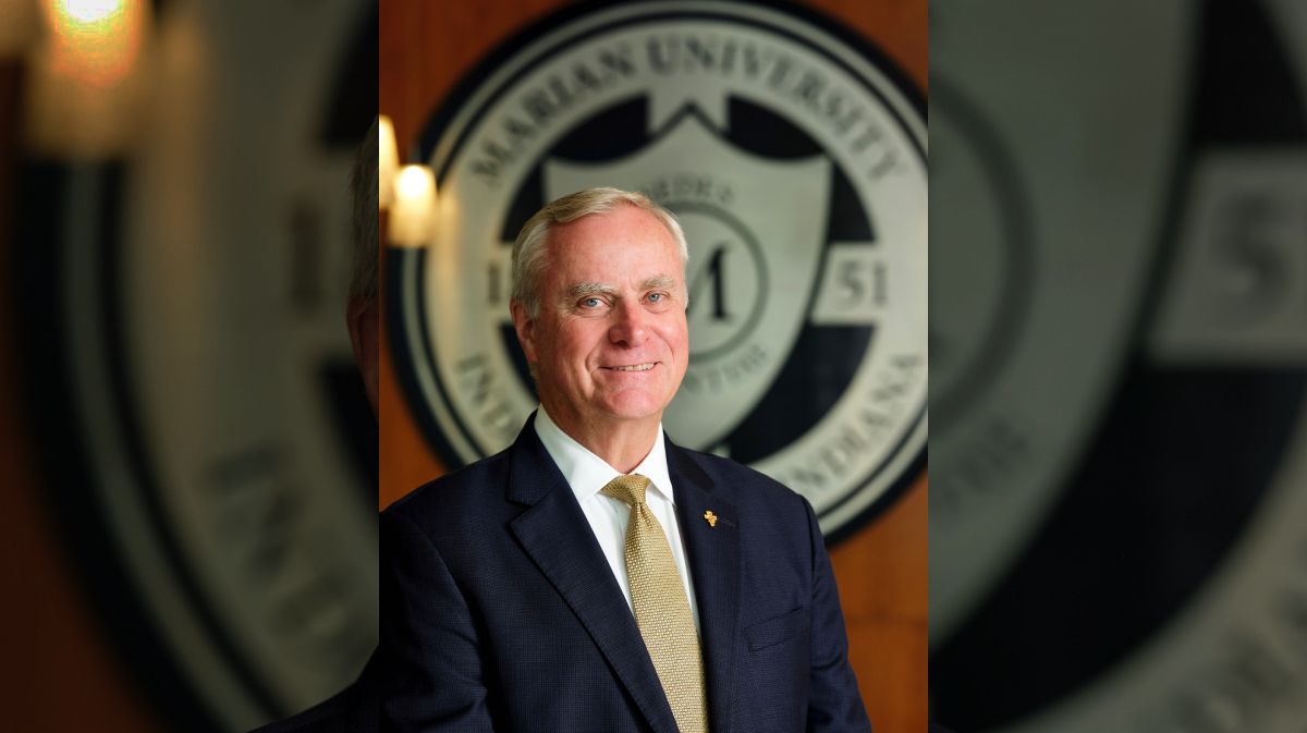 Portrait of a smiling older man in a dark suit and gold tie, standing in front of a large blurred university seal.