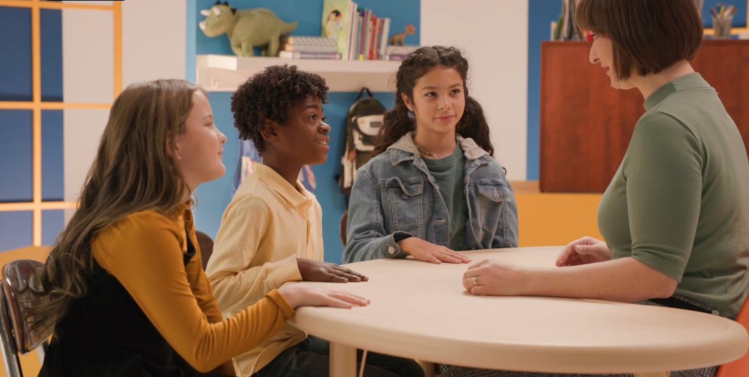 Teacher talking with three students at a round table in a classroom.