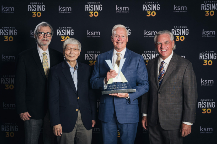 Four men in suits pose at a Conexus Rising 30 event backdrop, with the man in the center holding a glass award.