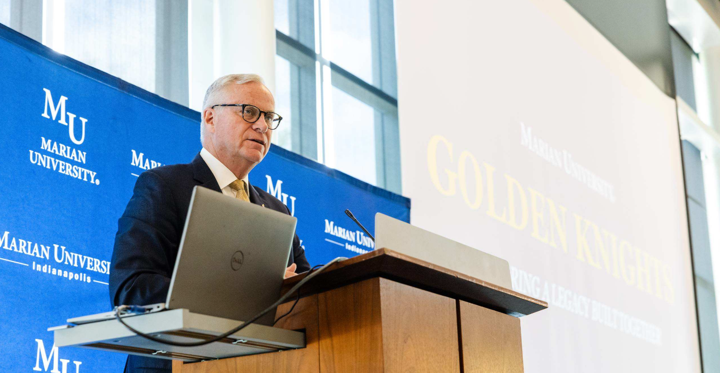 Speaker at a podium with a laptop, in front of a Marian University backdrop and a projection screen.