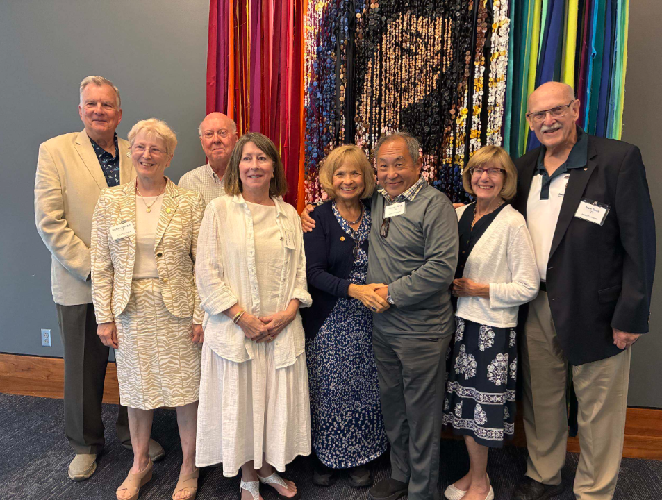 Group of eight adults posing together at an indoor event in front of a colorful ribbon-and-sequin wall hanging.