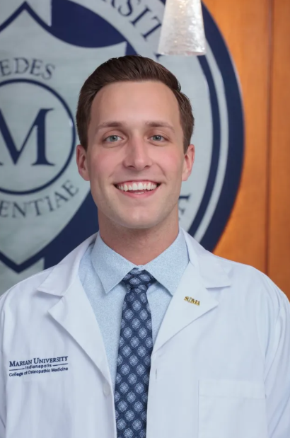 Smiling man in a white lab coat and tie posing for a formal portrait in front of a university emblem.