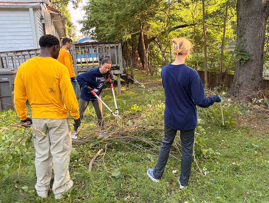 Volunteers clear brush and branches in a grassy backyard beside a house and deck, using loppers and gloves on a sunny day.