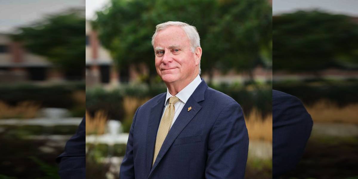 Older man in a navy suit and gold tie posing outdoors with blurred greenery in the background.