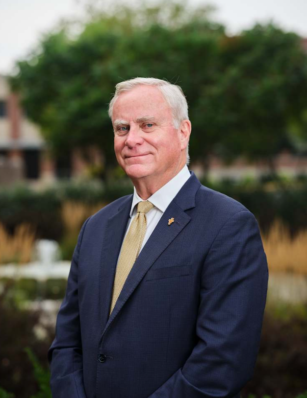 Older man in a navy suit and gold tie posing outdoors with blurred greenery in the background.