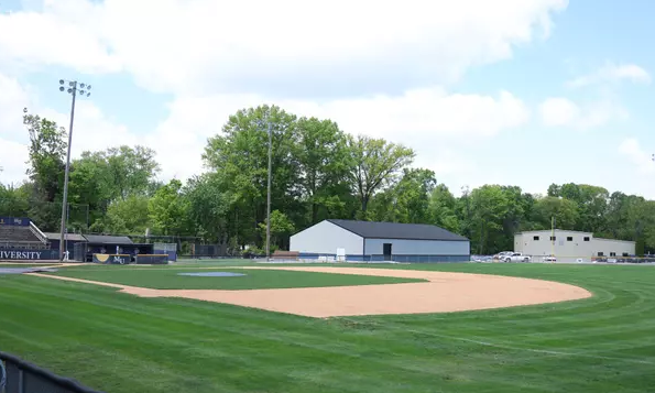 Empty baseball field with a well-kept infield and green outfield, bordered by trees and small buildings under a partly cloudy sky.