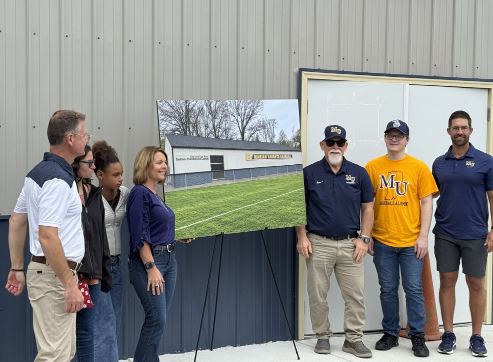Group of people pose by a building entrance next to a large poster board showing a baseball field facility rendering.