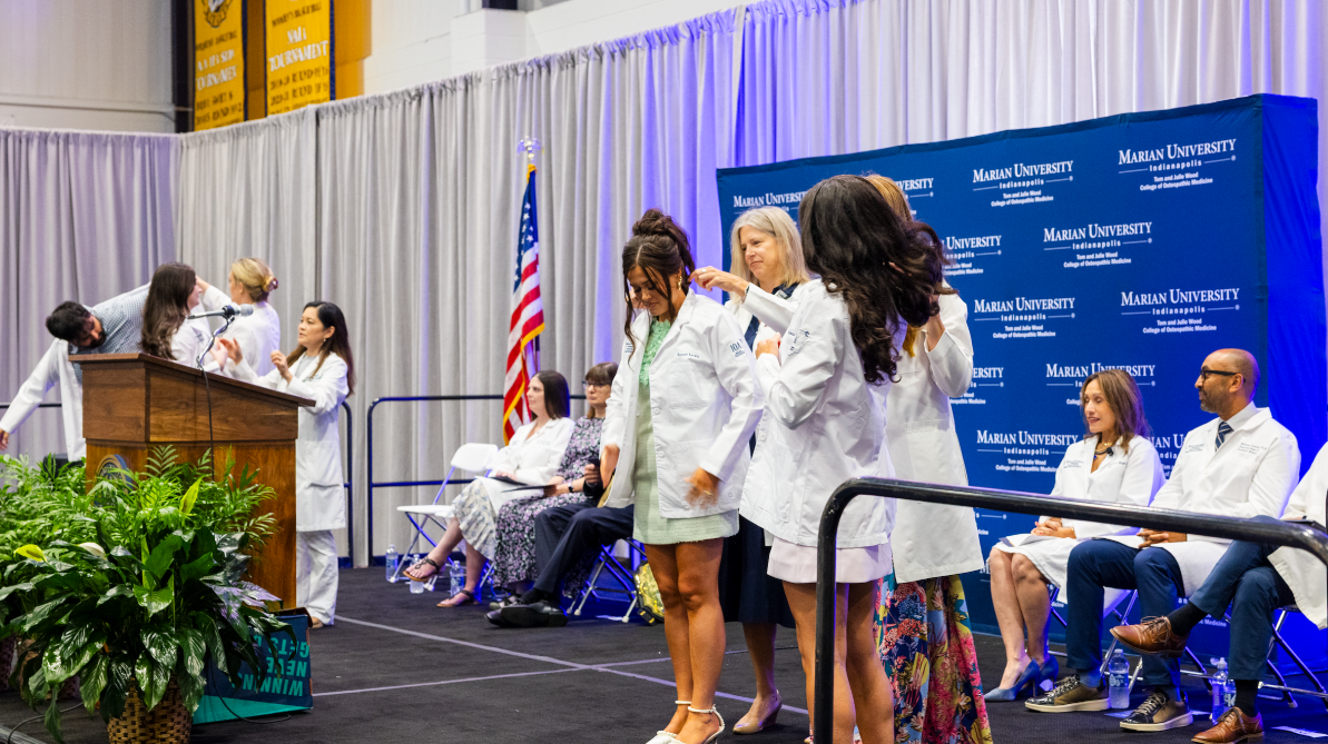 White-coat ceremony on a stage at Marian University, with students in white coats and faculty seated nearby as one student is helped into her coat.